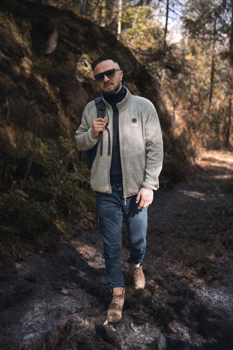 Person hiking through a forest trail wearing sunglasses, a gray jacket, jeans, and brown boots, carrying a backpack, with sunlight filtering through the trees.